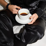 Person in black silk pajamas holding a white cup of coffee.