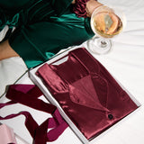 Burgundy silk pyjamas in christmas gift box, girl sits close by holding champagne wearing green silk