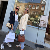 Two stylish women posing outside the Silk Works Chelsea pop-up, holding white branded gift bags in front of a floral window display and boutique signage.