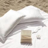 Silk Works white silk pillowcases, eye mask, and scrunchie styled on the beach with an open book, capturing a serene self-care moment.