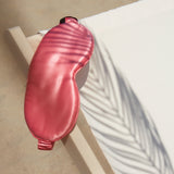 Silk Works watermelon pink mulberry silk eye mask resting on a deck chair with palm leaf shadows, styled in a sun-drenched beach setting.