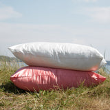 Stack of Silk Works mulberry silk pillowcases in ivory and watermelon pink, styled outdoors on grass under a bright summer sky.