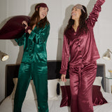 Two women in silk pyjamas on a bed, one in green and one in maroon.