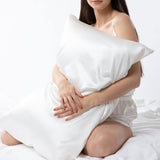 Woman sitting on a bed holding a large ivory Silk Works mulberry silk pillowcase, dressed in ivory cami sleep set