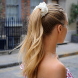 Woman wearing a high ponytail secured with a Silk Works ivory silk scrunchie, walking outdoors in a soft pink off-shoulder top.