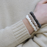 Set of three skinny silk scrunchies in caramel, ivory, and charcoal grey worn on a model’s wrist – showcasing their soft texture and neutral tones