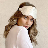 Woman wearing a Silk Works ivory mulberry silk eye mask with ruched strap, styled with soft waves and a white top, against a neutral textured background.