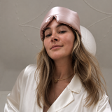 Woman wearing a pink silk large eye mask and white silk pyjams against a neutral background