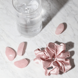 Pink silk scrunchie and rose quartz crystals on a marble surface with a glass of water.