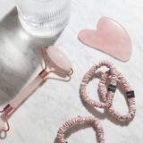 Pink skincare tools including a guasha, and hair ties on a marble surface with a glass of water.