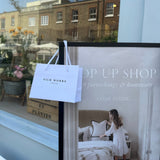 Silk Works branded gift bag hanging on a pop-up shop sign, with a lifestyle poster inviting guests to explore soft furnishings and silk homeware inside.