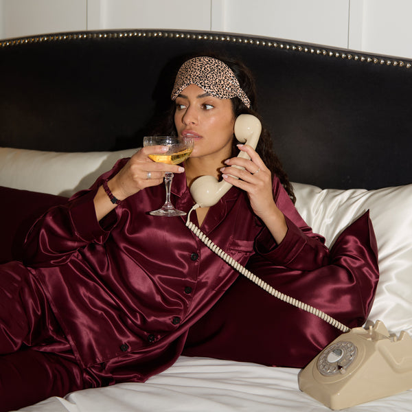 Woman in burgundy silk pyjamas lying on a bed, holding a glass of wine and talking on a vintage phone.